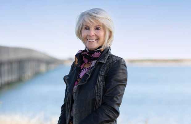 Participant Joanne - person with short white hair wearing a colorful patterned scarf and smiling in front of a beach with a bridge
