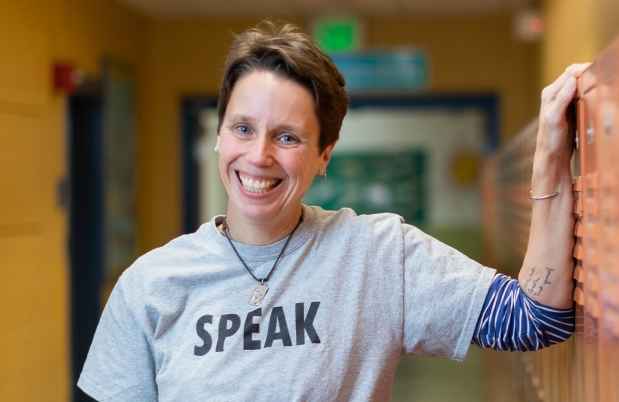 Participant Jen - person with short auburn hair wearing a grey shirt that says speak leaning against orange lockers in a hallway