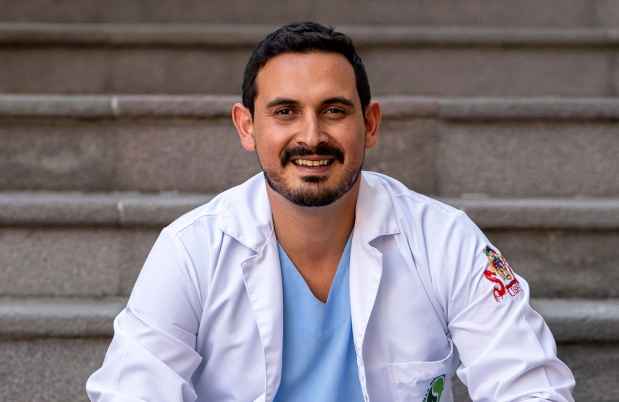 Deconstructing Stigma participant Andres - person in lab coat and scrubs sits on outdoor steps