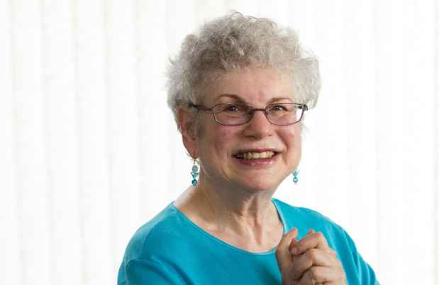 Participant Carol - person with short white hair wearing teal and smiling in front of a bright white background