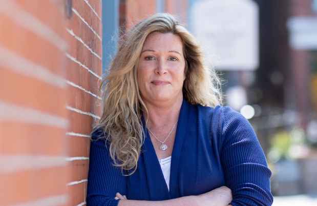 Participant Jenny - person with long blond hair wearing a blue cardigan and leaning against a brick wall