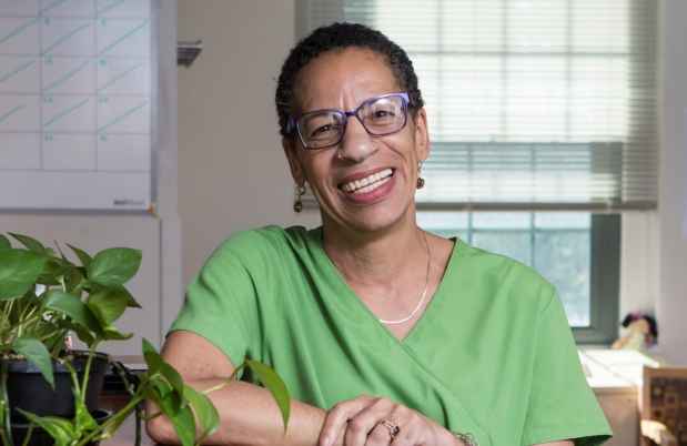 Participant Joyce - person with short dark hair wearing glasses and green medical scrubs smiles by a table inside