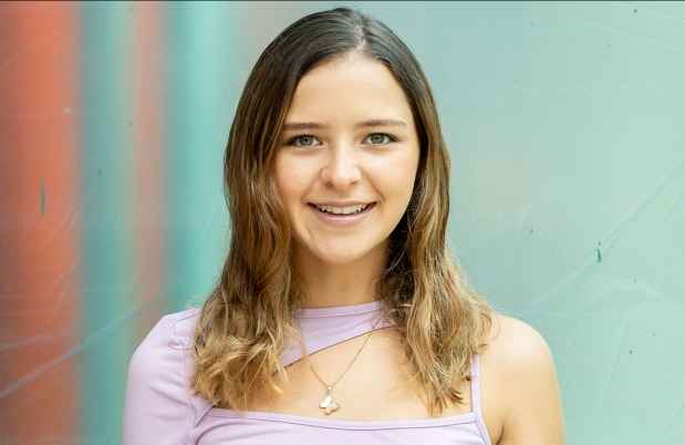 Participant Alejandra - person with brown wavy hair and a purple shirt smiling in front of a bright teal wall