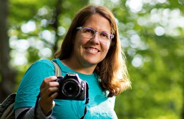 Participant Marlena - person with brown hair and glasses posing with a camera in a forest