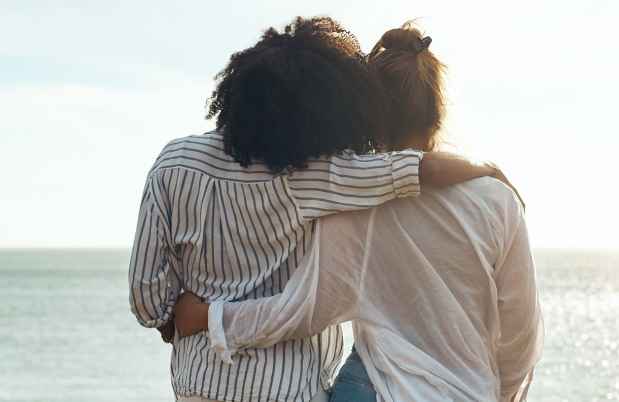 Two people with arms around each other looking at the ocean