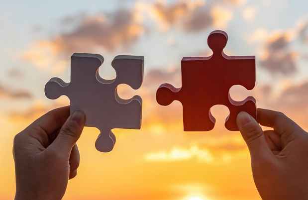 Two hands holding large jigsaw puzzle pieces in front of a sunset