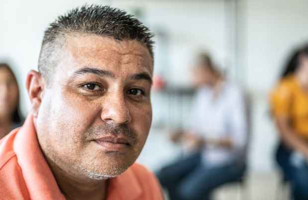 Portrait of a person with short hair wearing a coral shirt. There are other people in the background