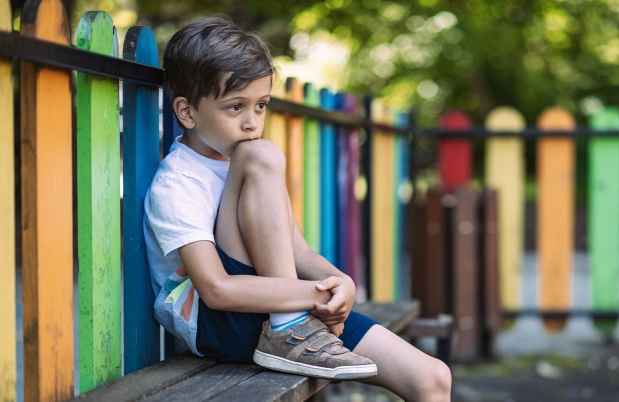 Young child sitting with a knee up on a bench in front of a colored wooded fence