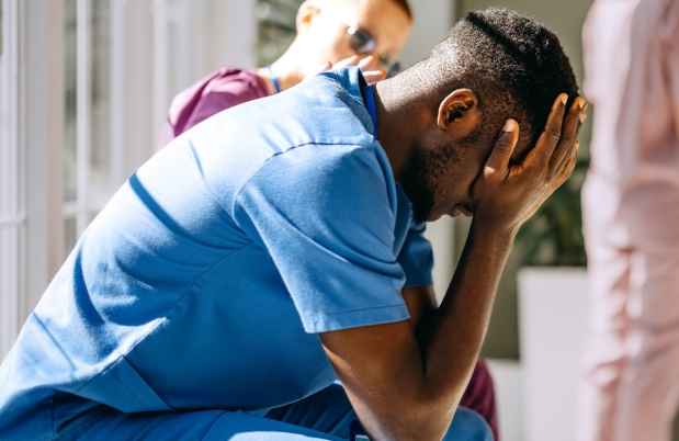 Man in scrubs sits holding his head in his hands