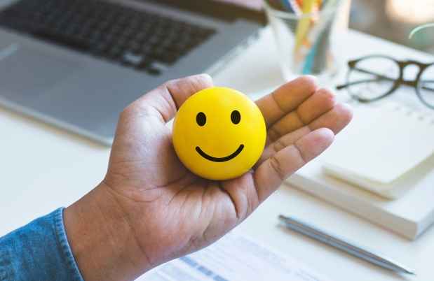 Hand holding a yellow ball with a smiley face on it. There is a desk with supplies like a laptop and glasses in the background