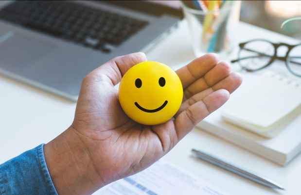Hand holding a yellow ball with a smiley face on it. There is a desk with supplies like a laptop and glasses in the background
