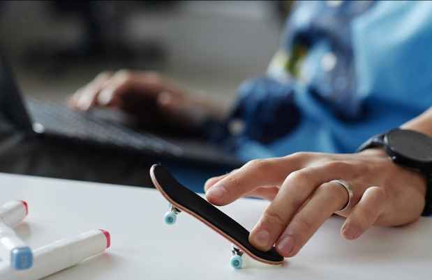 Person sitting with a laptop on their lap using their fingers to play with a tiny skateboard on the desk in front of them
