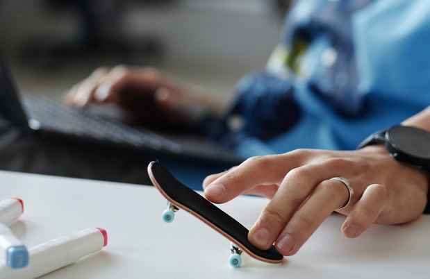 Person sitting with a laptop on their lap using their fingers to play with a tiny skateboard on the desk in front of them