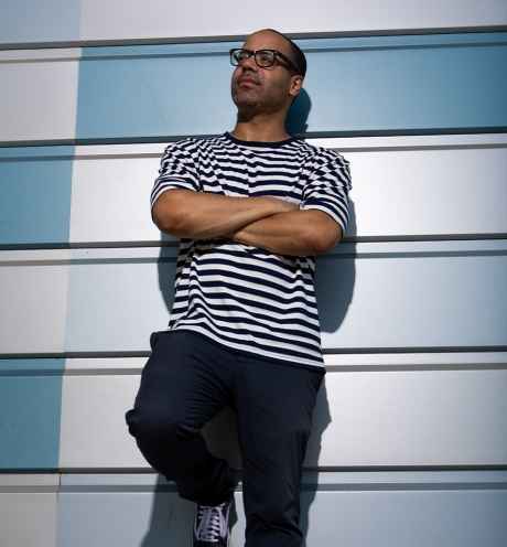 Participant Richard - person with short hair and glasses in a striped shirt leaning against a blue geometric wall