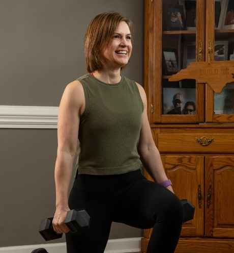Participant Stephanie - person with short brown hair wearing a green tank top smiling and lunging holding weights