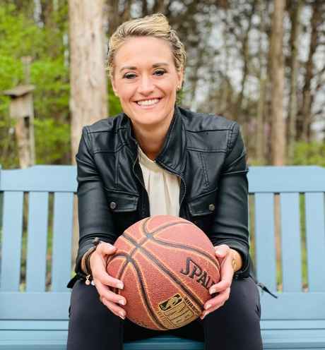 Participant Mandy - person with blond hair pulled back into an updo wearing a black leather jacket, sitting on a blue bench and holding a basketball