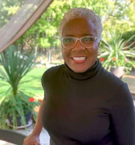 Participant Marilyn - person with short grey hair wearing a black turtleneck and smiling in front of plants