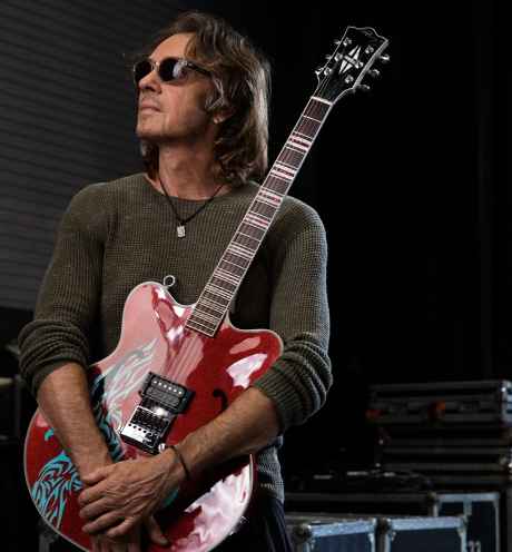 Participant Rick - person with long hair and sunglasses in front of a dark background holding a guitar