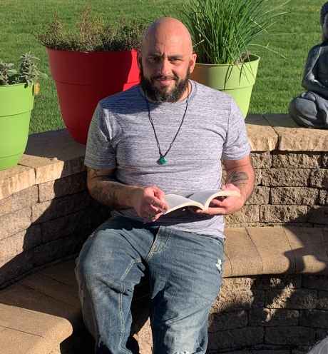Participant Stephen - person with dark facial hair smiling holding a book in front of grass and potted plants