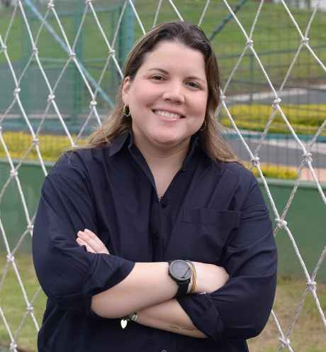 Participant Sylvia - person with long dark hair smiling with crossed arms in front of a fence outside