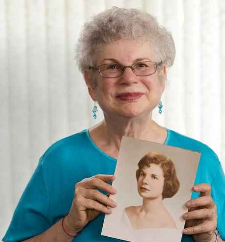 Participant Carol - person with short white hair wearing teal, smiling in front of a bright white background and holding a framed illustration