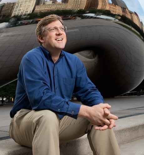 Participant John - person with short brown hair and glasses smiling in front of a large mirrored surface 