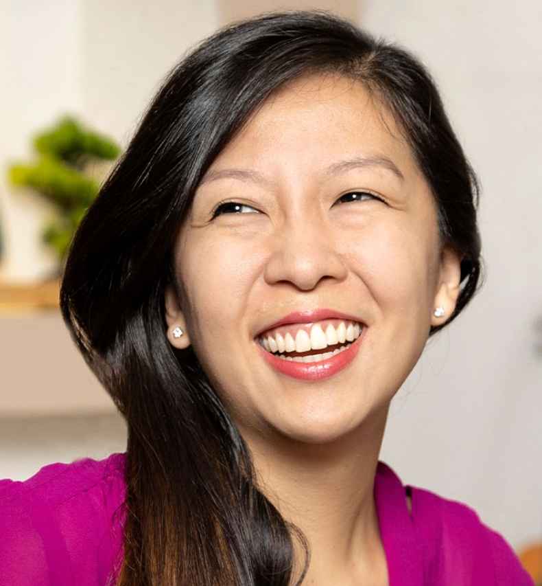 Participant Lena - person with long dark hair in a pink blouse smiling in front of a bookshelf