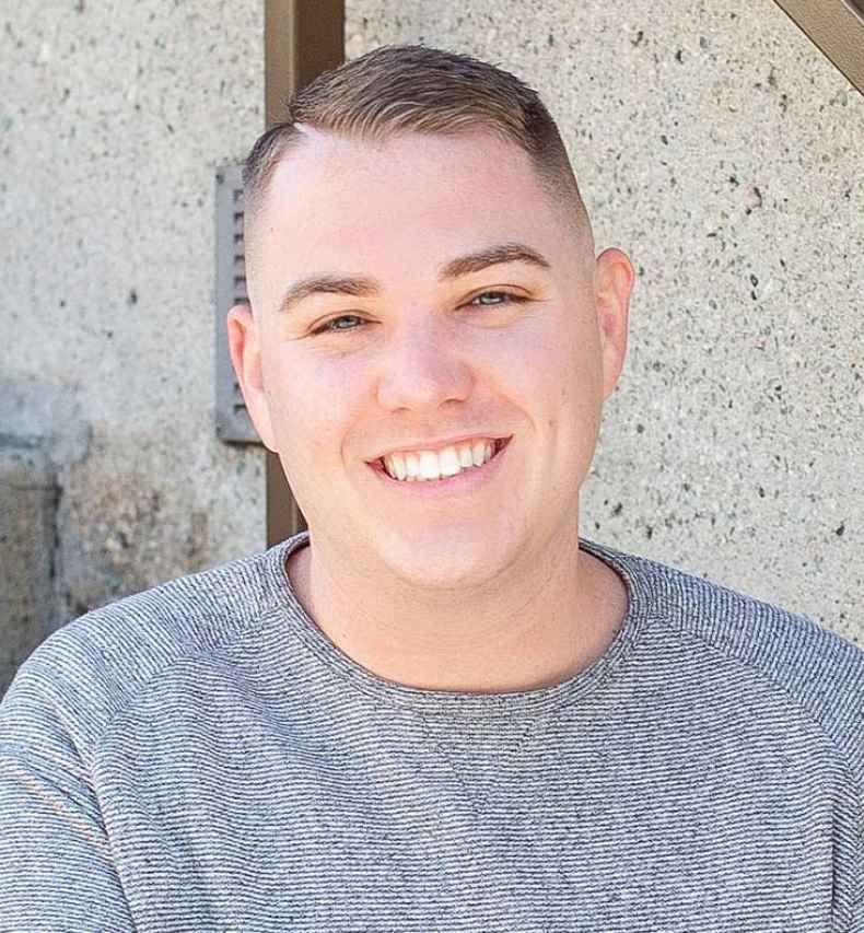 Participant Kyle - person with short brown hair in a grey sweater smiling and sitting on concrete steps