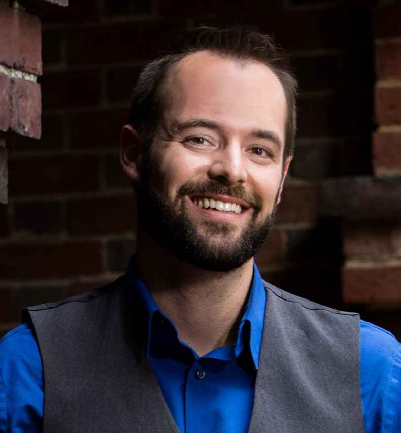 Participant Nathaniel - person with short dark hair wearing a blue shirt leaning against a brick pillar