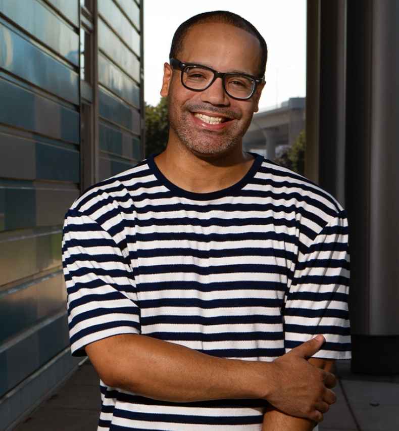 Participant Richard - person with short hair and glasses in a striped shirt smiling in front of a blue geometric wall