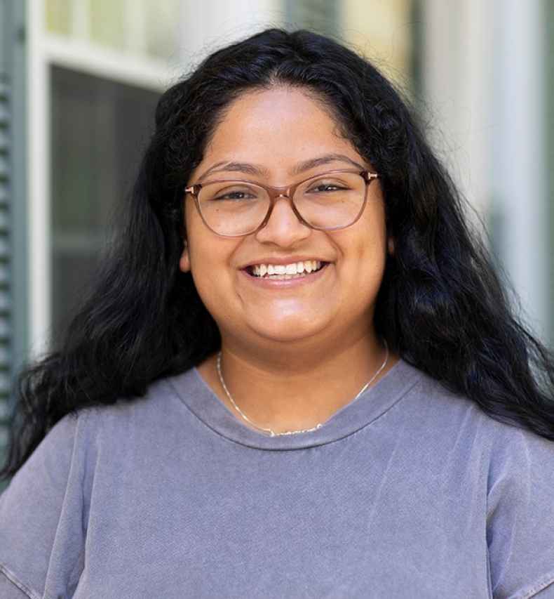 Participant Marina - person with dark hair and glasses wearing a grey t-shirt smiling in front of a building