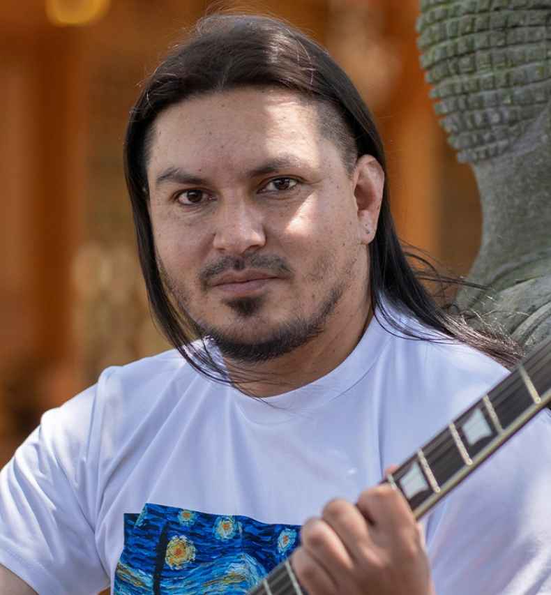 Participant Pablo - person with long dark hair holding a guitar and leaning against a statue