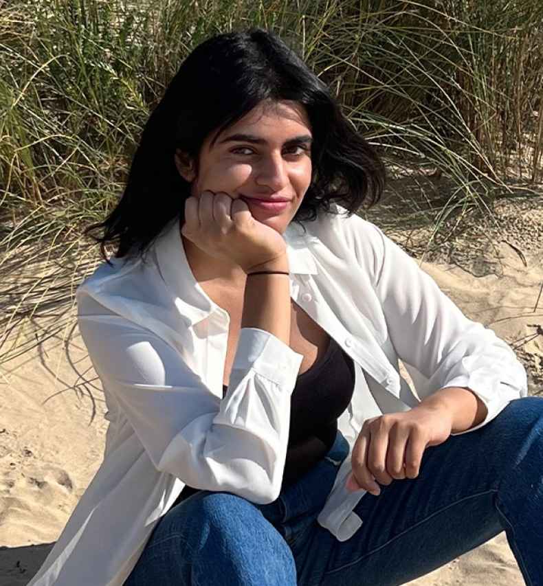 Participant Samina - person wearing jeans and a white coat sitting on sand at a beach