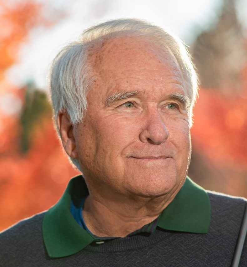 Participant Mike - person with short white hair holding a golf club in front of fall foliage