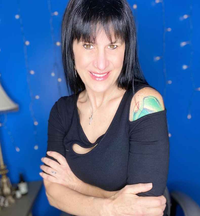 Participant Nique - person with long dark hair smiling with crossed arms in front of a blue wall
