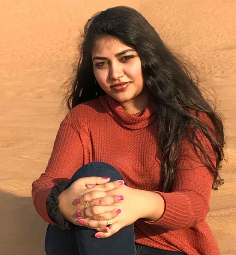 Participant Ramya - person with long dark hair in a red sweater sitting in sand