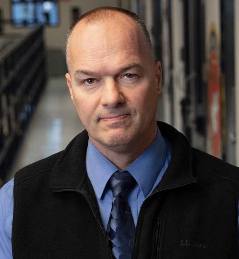 Participant Randall - a person wearing a blue shirt and a black vest in front of a hallway