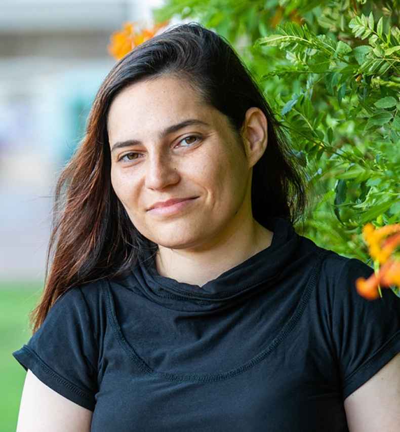 Deconstructing Stigma participant Shelly - Person in black shirt and long hair stands outside with greenery behind