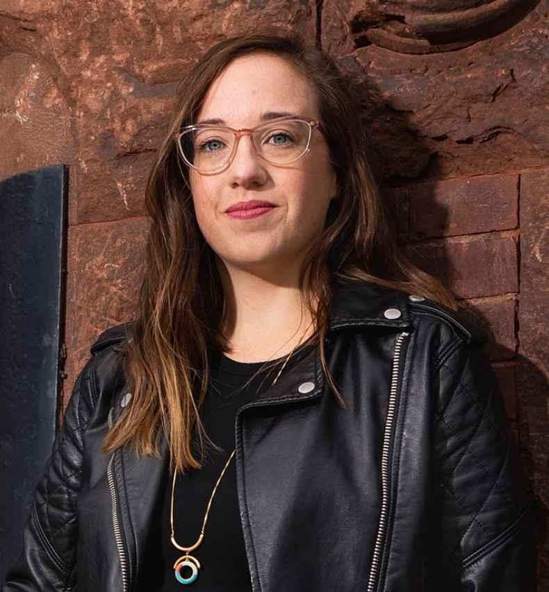 Participant Angi - person with long wavy hair and glasses wearing a black leather jacket and leaning against a stone building