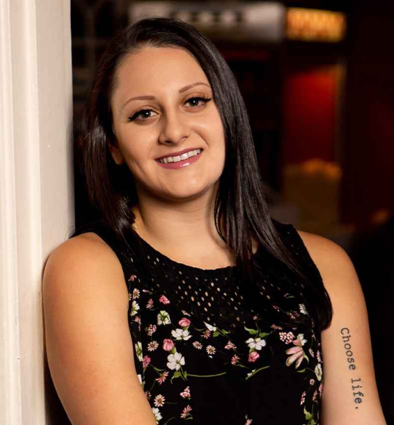 Participant Jackie - person with long dark hair smiling and leaning against a wall