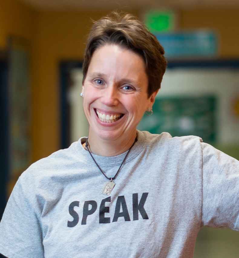 Participant Jen - person with short auburn hair wearing a grey shirt that says speak leaning against orange lockers in a hallway