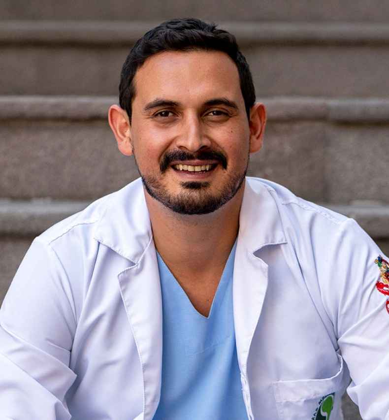 Deconstructing Stigma participant Andres - person in lab coat and scrubs sits on outdoor steps