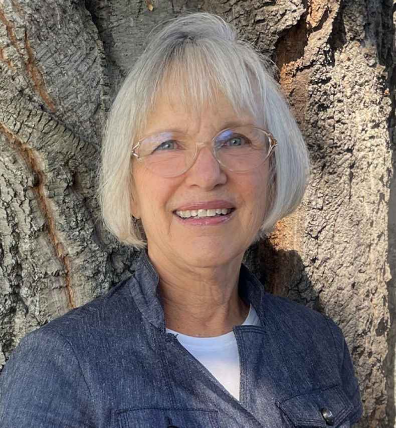 Participant Barbara - person with short white hair smiling in front of a tree trunk