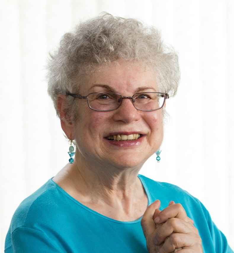 Participant Carol - person with short white hair wearing teal and smiling in front of a bright white background