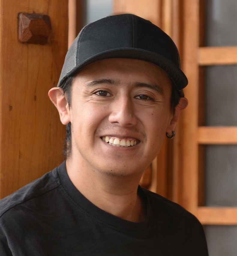 Participant Jimmy - person with short dark hair wearing a black baseball hat and smiling outside a wooden building