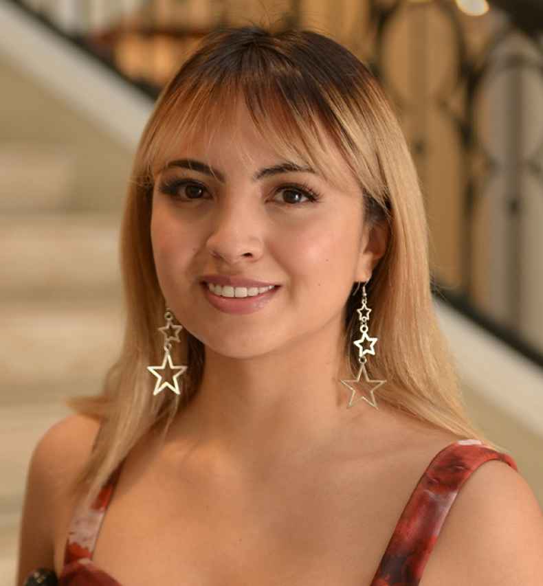 Participant Valeria - person with brown hair wearing star-shaped earrings and a red tank top smiling near a set of stone stairs