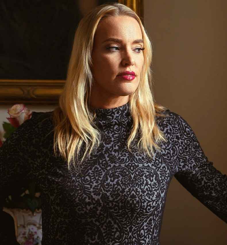 Participant Elsie - person with long blond hair wearing a dress in front of a painting and flowers