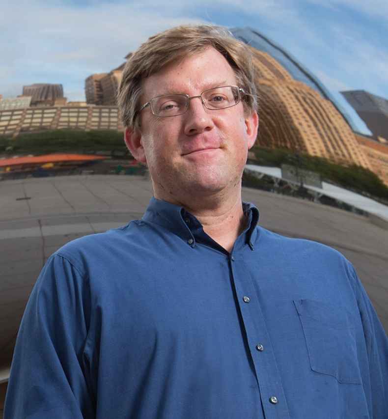 Participant John - person with short brown hair and glasses smiling in front of a large mirrored surface 