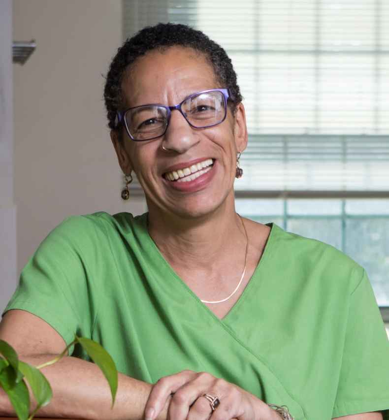 Participant Joyce - person with short dark hair wearing glasses and green medical scrubs smiles by a table inside