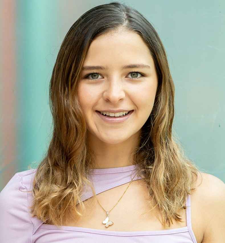 Participant Alejandra - person with brown wavy hair and a purple shirt smiling in front of a bright teal wall